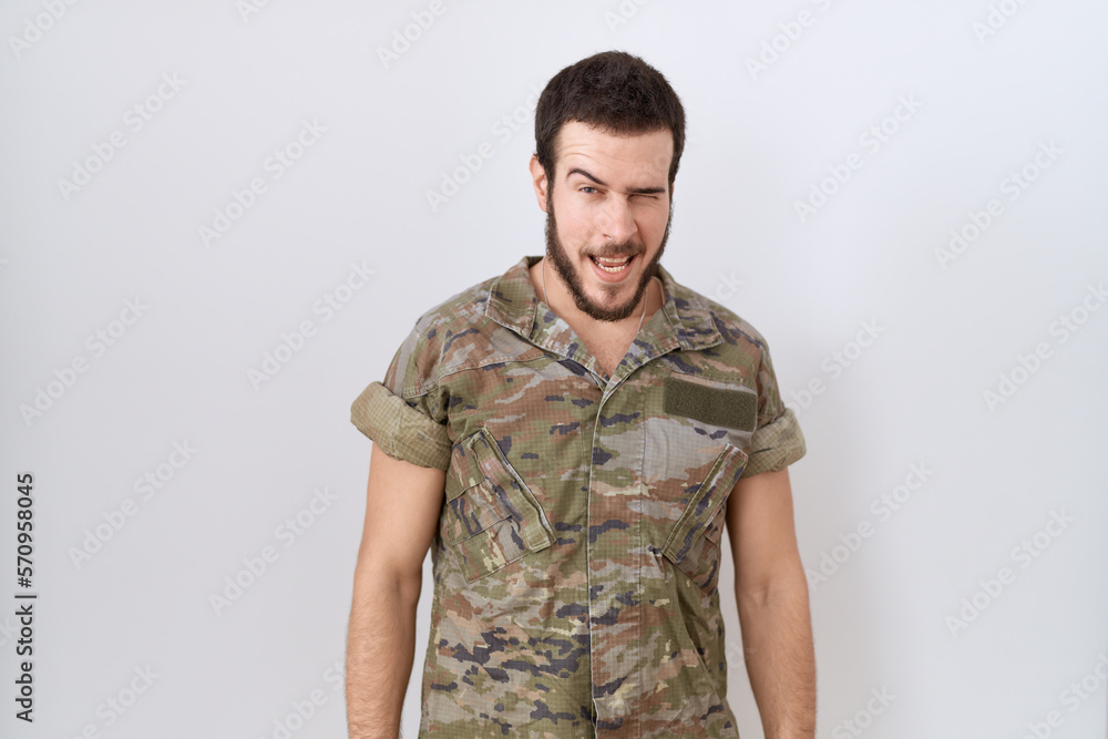 Young hispanic man wearing camouflage army uniform winking looking at the camera with sexy expression, cheerful and happy face. Young hispanic man wearing camouflage army uniform winking looking at the camera with sexy expression, cheerful and happy face.