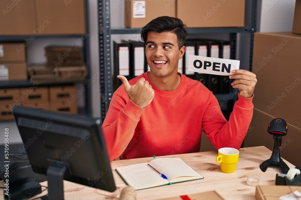 Hispanic man working at small business ecommerce holding open banner pointing thumb up to the side smiling happy with open mouth