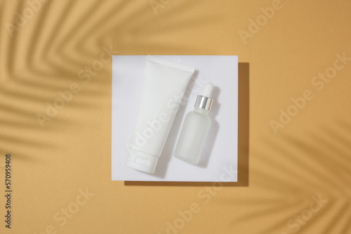 Skincare products concept. Top view photo of white cream tube, dropper bottle on square podium and tropical leaves shadow on sandy background. Summer cosmetics mockup idea.