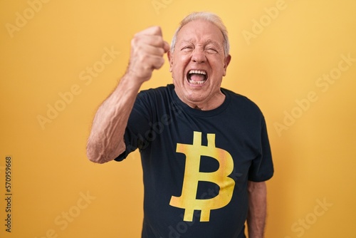 Senior man with grey hair wearing bitcoin t shirt angry and mad raising fist frustrated and furious while shouting with anger. rage and aggressive concept.