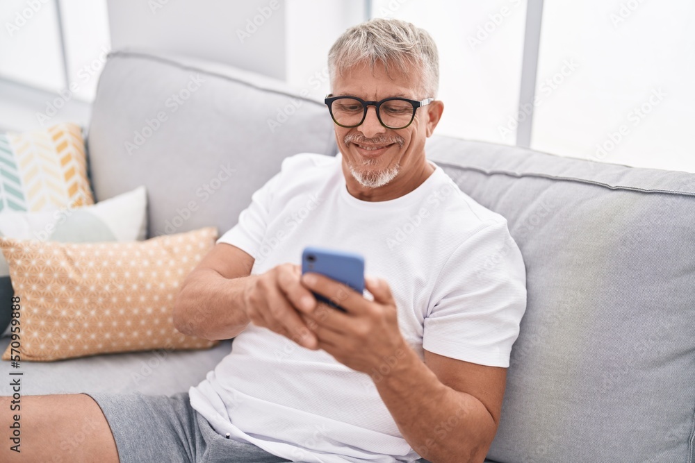 Middle age grey-haired man using smartphone sitting on sofa at home Middle age grey-haired man using smartphone sitting on sofa at home