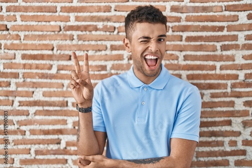 Brazilian young man standing over brick wall smiling with happy face winking at the camera doing victory sign with fingers. number two.