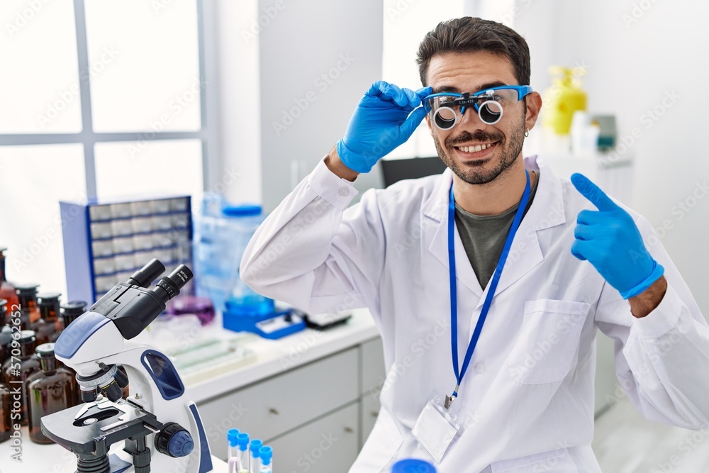 Young hispanic man working at scientist laboratory wearing magnifying glasses smiling happy pointing with hand and finger Young hispanic man working at scientist laboratory wearing magnifying glasses smiling happy pointing with hand and finger
