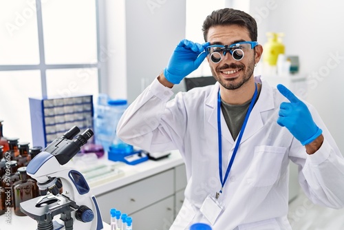 Young hispanic man working at scientist laboratory wearing magnifying glasses smiling happy pointing with hand and finger
