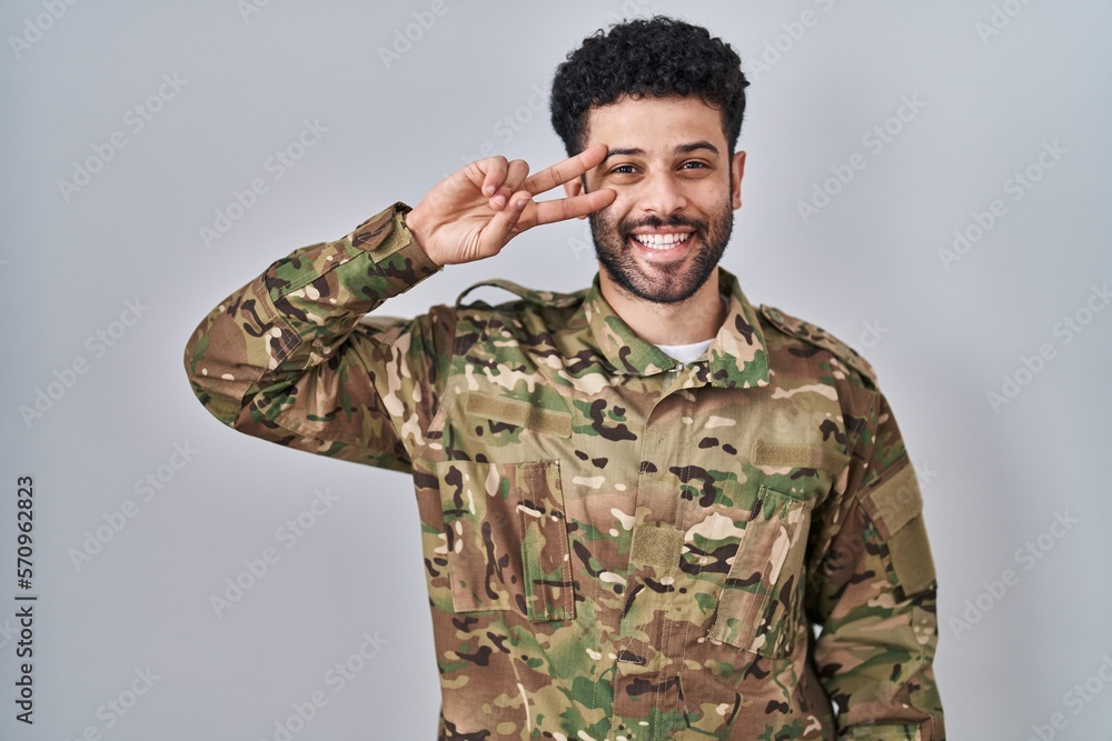 Arab man wearing camouflage army uniform doing peace symbol with fingers over face, smiling cheerful showing victory