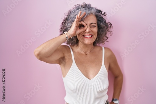 Middle age woman with grey hair standing over pink background doing ok gesture with hand smiling, eye looking through fingers with happy face.