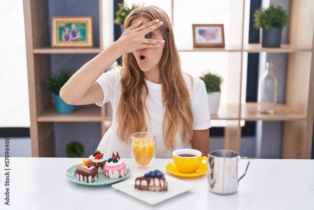 Young caucasian woman eating pastries t for breakfast peeking in shock covering face and eyes with hand, looking through fingers with embarrassed expression.