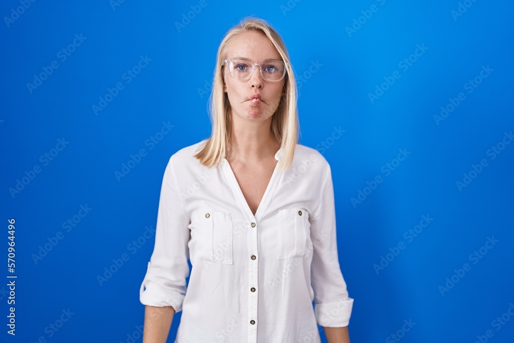 Young caucasian woman standing over blue background making fish face with lips, crazy and comical gesture. funny expression. Young caucasian woman standing over blue background making fish face with lips, crazy and comical gesture. funny expression.