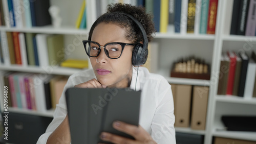 African american woman student using touchpad and headphones with doubt expression at library university
