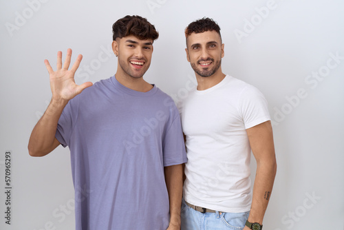 Homosexual gay couple standing over white background showing and pointing up with fingers number five while smiling confident and happy.