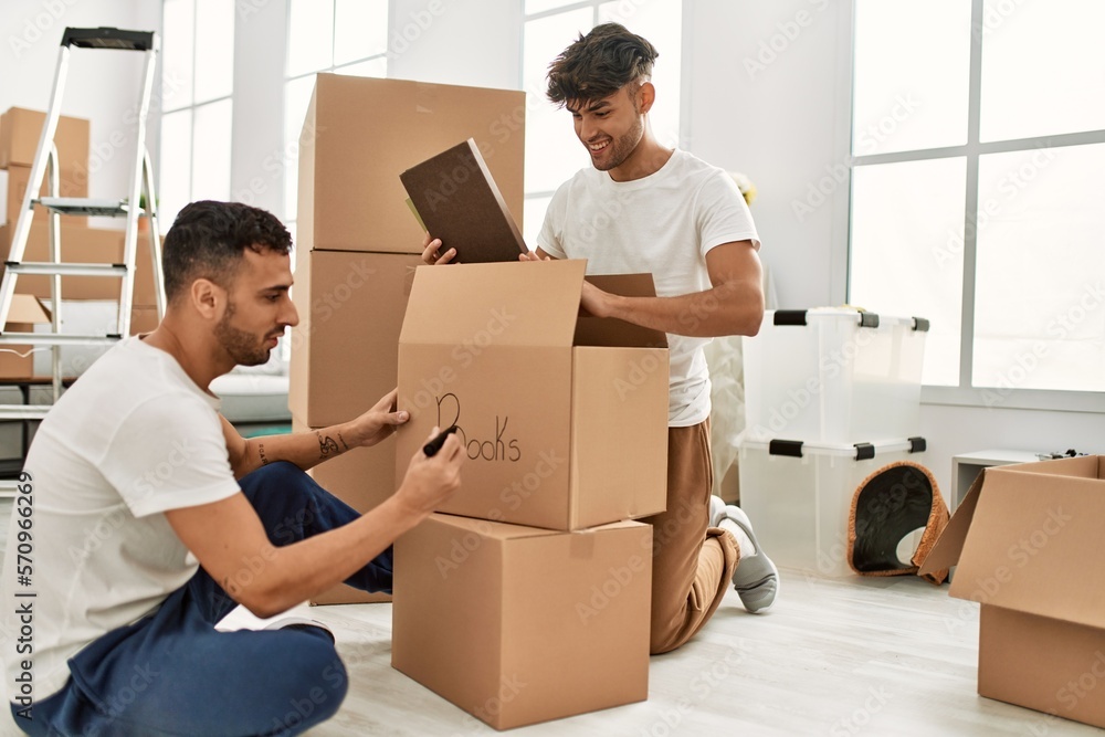 Two hispanic men couple smiling confident packing book cardboard box at new home Two hispanic men couple smiling confident packing book cardboard box at new home