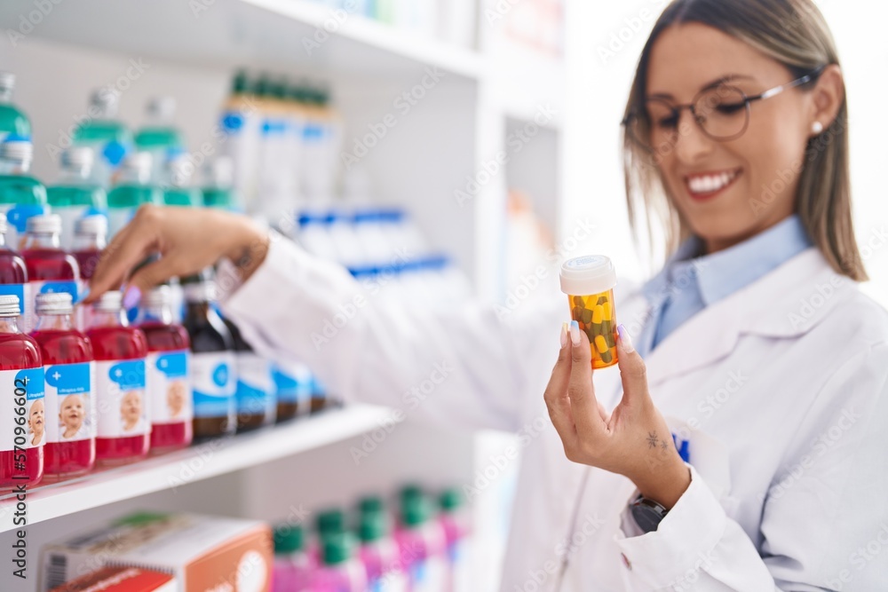 Young beautiful hispanic woman pharmacist smiling confident holding pills bottle at pharmacy