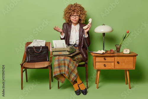 Confused curly haired elegant lady doesnt know telephone number holds handset poses with typewriter on knees wears old fashionable clothing isolated over green background. Retro style concept © wayhome.studio 