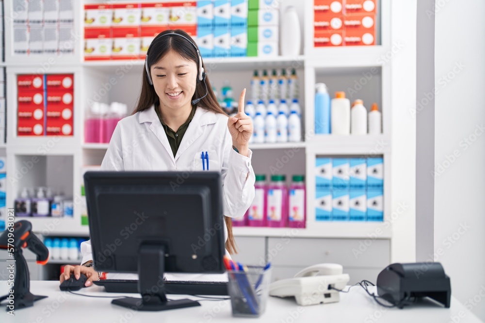 Chinese young woman working at pharmacy drugstore wearing headset smiling with an idea or question pointing finger with happy face, number one Chinese young woman working at pharmacy drugstore wearing headset smiling with an idea or question pointing finger with happy face, number one