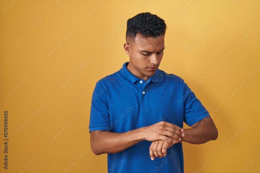 Young hispanic man standing over yellow background checking the time on wrist watch, relaxed and confident Young hispanic man standing over yellow background checking the time on wrist watch, relaxed and confident