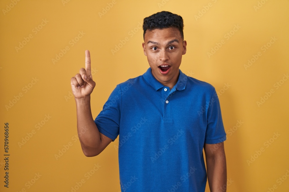 Young hispanic man standing over yellow background pointing finger up with successful idea. exited and happy. number one. Young hispanic man standing over yellow background pointing finger up with successful idea. exited and happy. number one.