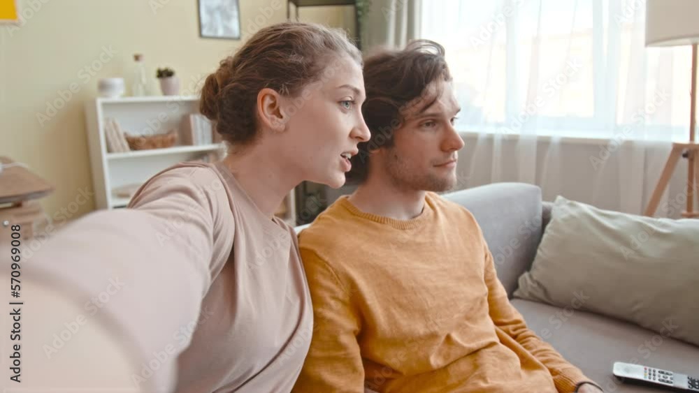Handheld POV shot of young Caucasian woman recording video of herself staying at home with boyfriend playing video games on console, sitting together on couch in living room Handheld POV shot of young Caucasian woman recording video of herself staying at home with boyfriend playing video games on console, sitting together on couch in living room