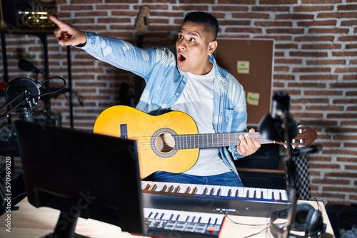 Hispanic young man playing classic guitar at music studio pointing with finger surprised ahead, open mouth amazed expression, something on the front