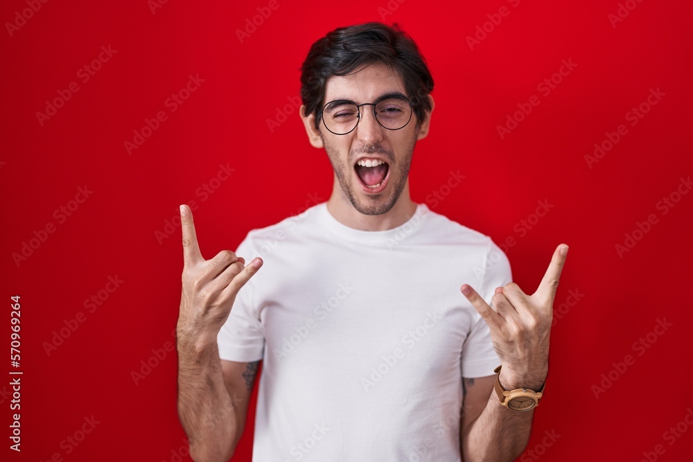 Naklejka premium Young hispanic man standing over red background shouting with crazy expression doing rock symbol with hands up. music star. heavy music concept.