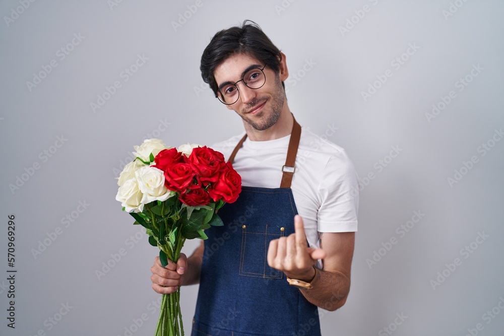 Young hispanic man holding bouquet of white and red roses beckoning come here gesture with hand inviting welcoming happy and smiling Young hispanic man holding bouquet of white and red roses beckoning come here gesture with hand inviting welcoming happy and smiling