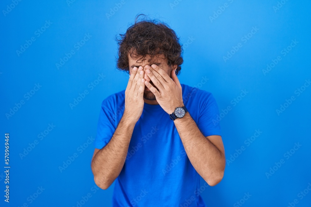 Hispanic young man standing over blue background rubbing eyes for fatigue and headache, sleepy and tired expression. vision problem Hispanic young man standing over blue background rubbing eyes for fatigue and headache, sleepy and tired expression. vision problem