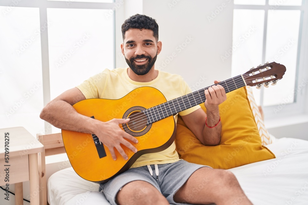 Young arab man playing classical guitar sitting on bed at bedroom