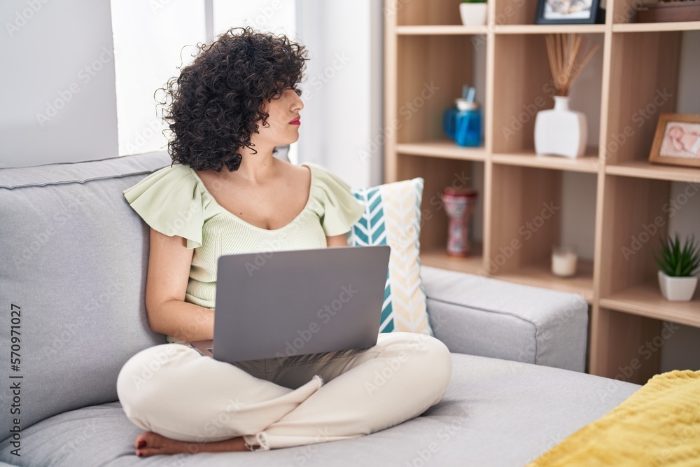 Young brunette woman with curly hair using laptop sitting on the sofa at home looking to side, relax profile pose with natural face with confident smile. Young brunette woman with curly hair using laptop sitting on the sofa at home looking to side, relax profile pose with natural face with confident smile.