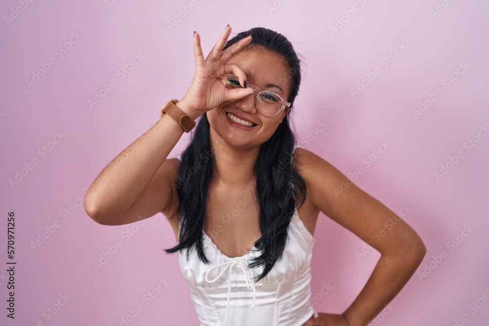 Asian young woman standing over pink background smiling happy doing ok sign with hand on eye looking through fingers Asian young woman standing over pink background smiling happy doing ok sign with hand on eye looking through fingers
