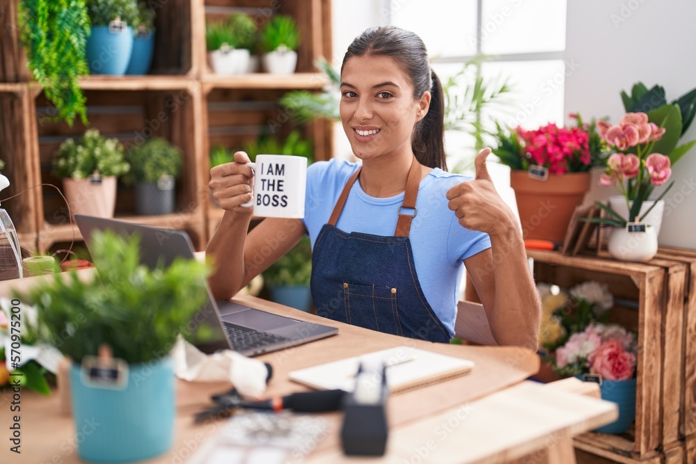 Brunette young woman working at florist shop holding i am the boss cup smiling happy and positive, thumb up doing excellent and approval sign Brunette young woman working at florist shop holding i am the boss cup smiling happy and positive, thumb up doing excellent and approval sign