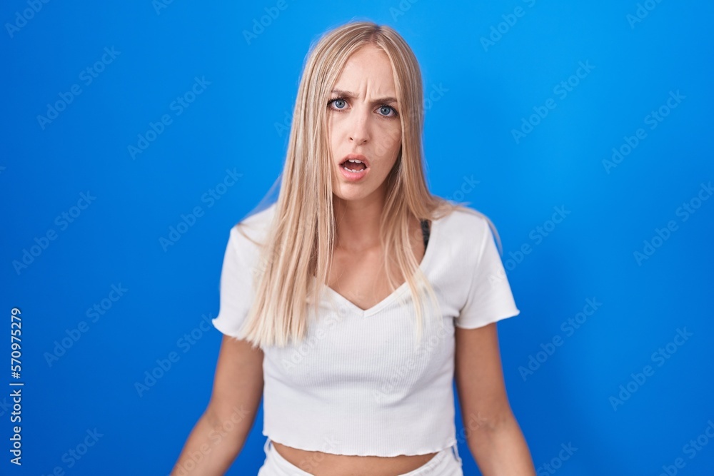 Young caucasian woman standing over blue background in shock face, looking skeptical and sarcastic, surprised with open mouth Young caucasian woman standing over blue background in shock face, looking skeptical and sarcastic, surprised with open mouth