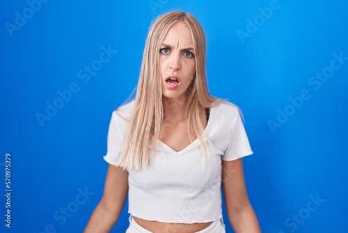 Young caucasian woman standing over blue background in shock face, looking skeptical and sarcastic, surprised with open mouth