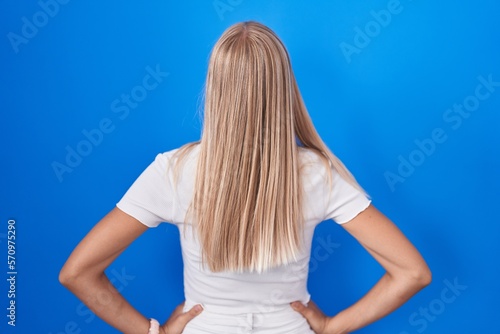 Young caucasian woman standing over blue background standing backwards looking away with arms on body