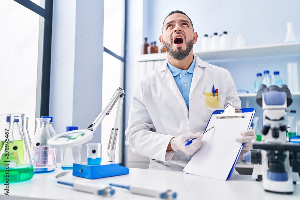 Hispanic man working at scientist laboratory holding blank clipboard angry and mad screaming frustrated and furious, shouting with anger looking up. Hispanic man working at scientist laboratory holding blank clipboard angry and mad screaming frustrated and furious, shouting with anger looking up.