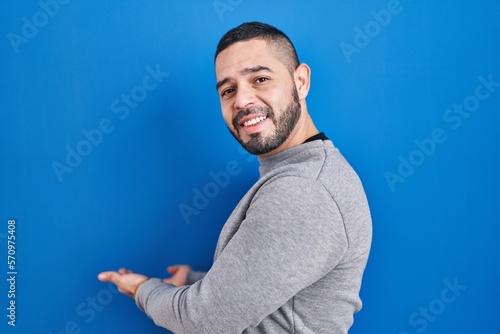 Hispanic man standing over blue background inviting to enter smiling natural with open hand