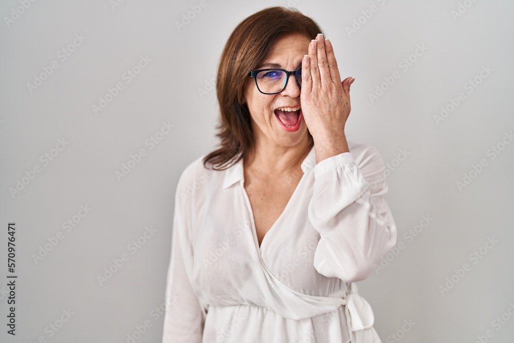 Middle age hispanic woman standing over isolated background covering one eye with hand, confident smile on face and surprise emotion. Middle age hispanic woman standing over isolated background covering one eye with hand, confident smile on face and surprise emotion.