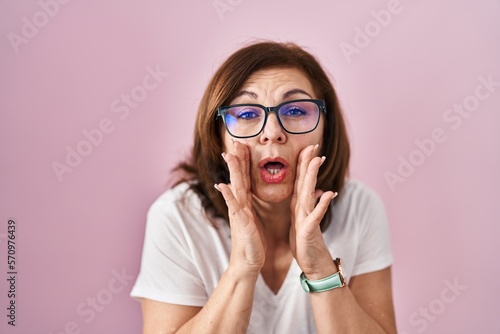 Middle age hispanic woman standing over pink background hand on mouth telling secret rumor, whispering malicious talk conversation