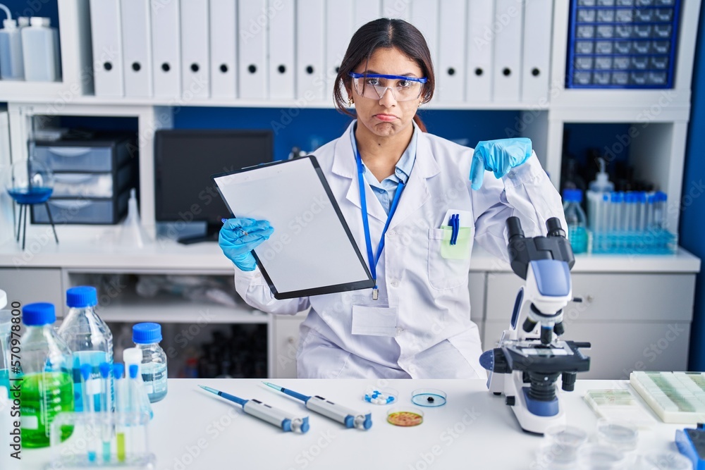 Hispanic young woman working at scientist laboratory pointing down looking sad and upset, indicating direction with fingers, unhappy and depressed. Hispanic young woman working at scientist laboratory pointing down looking sad and upset, indicating direction with fingers, unhappy and depressed.