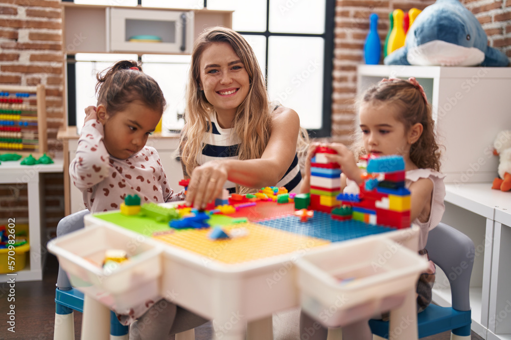 Fototapeta premium Teacher with girls playing with construction blocks sitting on table at kindergarten