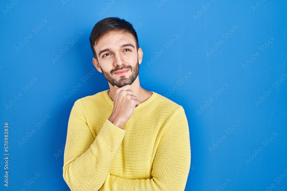 Hispanic man standing over blue background with hand on chin thinking about question, pensive expression. smiling and thoughtful face. doubt concept. Hispanic man standing over blue background with hand on chin thinking about question, pensive expression. smiling and thoughtful face. doubt concept.