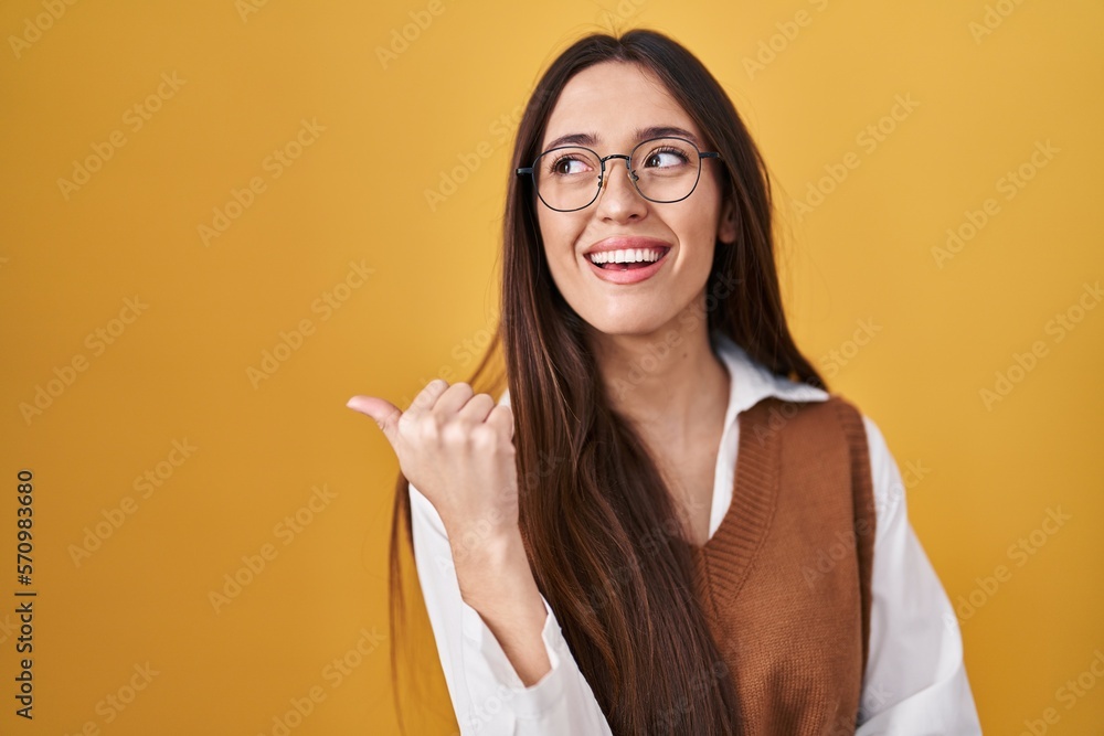Young brunette woman standing over yellow background wearing glasses smiling with happy face looking and pointing to the side with thumb up.