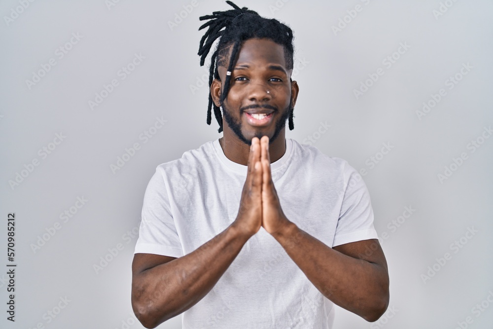 African man with dreadlocks wearing casual t shirt over white background praying with hands together asking for forgiveness smiling confident. African man with dreadlocks wearing casual t shirt over white background praying with hands together asking for forgiveness smiling confident.