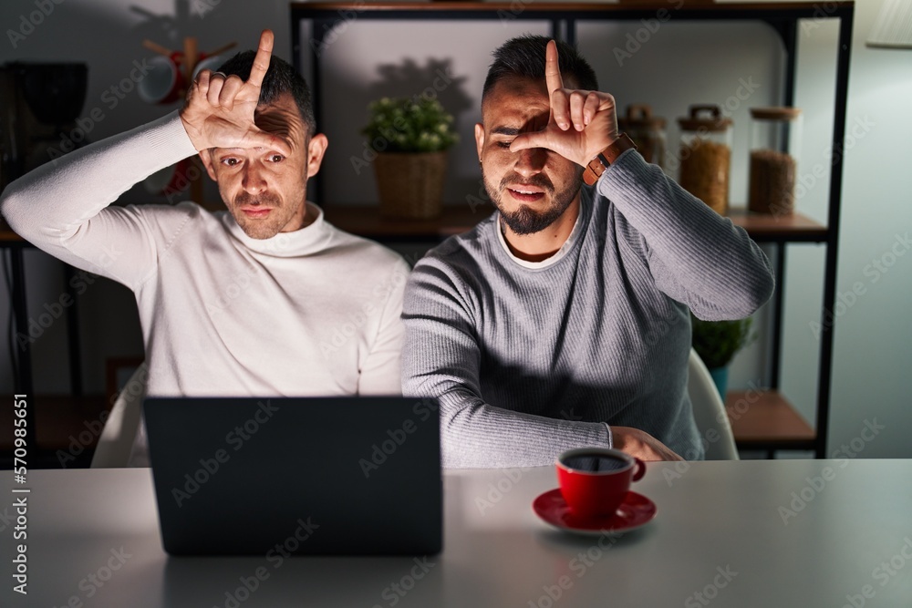 Homosexual couple using computer laptop making fun of people with fingers on forehead doing loser gesture mocking and insulting. Homosexual couple using computer laptop making fun of people with fingers on forehead doing loser gesture mocking and insulting.