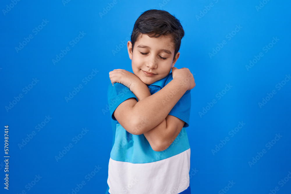 Little hispanic boy standing over blue background hugging oneself happy and positive, smiling confident. self love and self care Little hispanic boy standing over blue background hugging oneself happy and positive, smiling confident. self love and self care