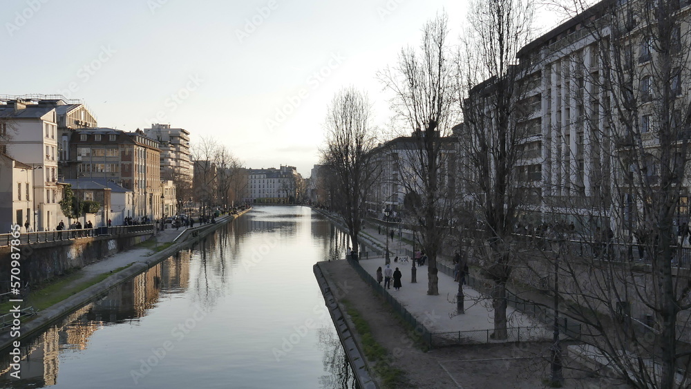 Canal Saint-Martin de Paris, pendant un couché de soleil, début de soirée, sombre, avec les immeubles peu éclairés par le Soleil et animé par un petit monde ou peu de gens, tranquille et paisible