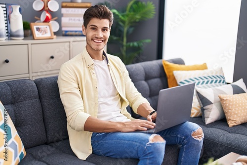 Young hispanic man using laptop sitting on sofa at home