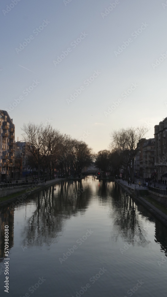 Canal Saint-Martin de Paris, pendant un couché de soleil, début de soirée, sombre, avec les immeubles peu éclairés par le Soleil et animé par un petit monde ou peu de gens, tranquille et paisible