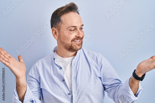 Middle age caucasian man standing over blue background smiling showing both hands open palms, presenting and advertising comparison and balance