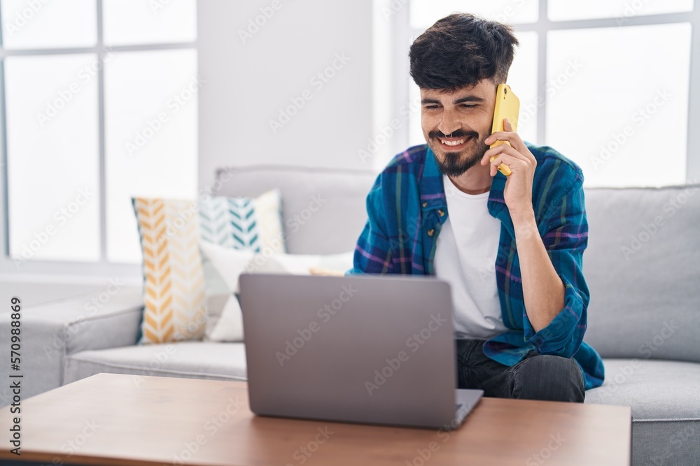 Young hispanic man using laptop and talking on smartphone sitting on sofa at home Young hispanic man using laptop and talking on smartphone sitting on sofa at home