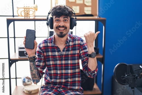 Young hispanic man with beard showing smartphone screen at music studio pointing thumb up to the side smiling happy with open mouth