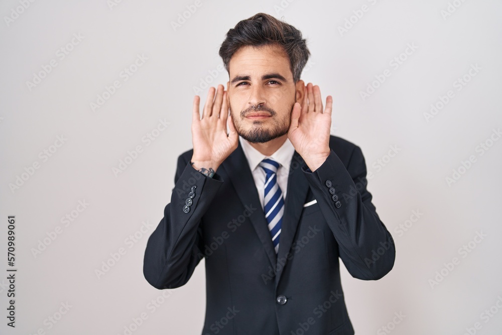 Young hispanic man with tattoos wearing business suit and tie trying to hear both hands on ear gesture, curious for gossip. hearing problem, deaf Young hispanic man with tattoos wearing business suit and tie trying to hear both hands on ear gesture, curious for gossip. hearing problem, deaf